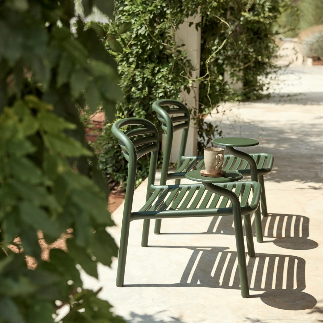 Green metal chairs with slatted design, casting playful shadows on a sunlit patio surrounded by lush greenery.