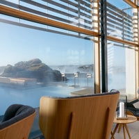 Natural wood tones with soft gray upholstery, featuring a calming blue landscape through large glass windows.