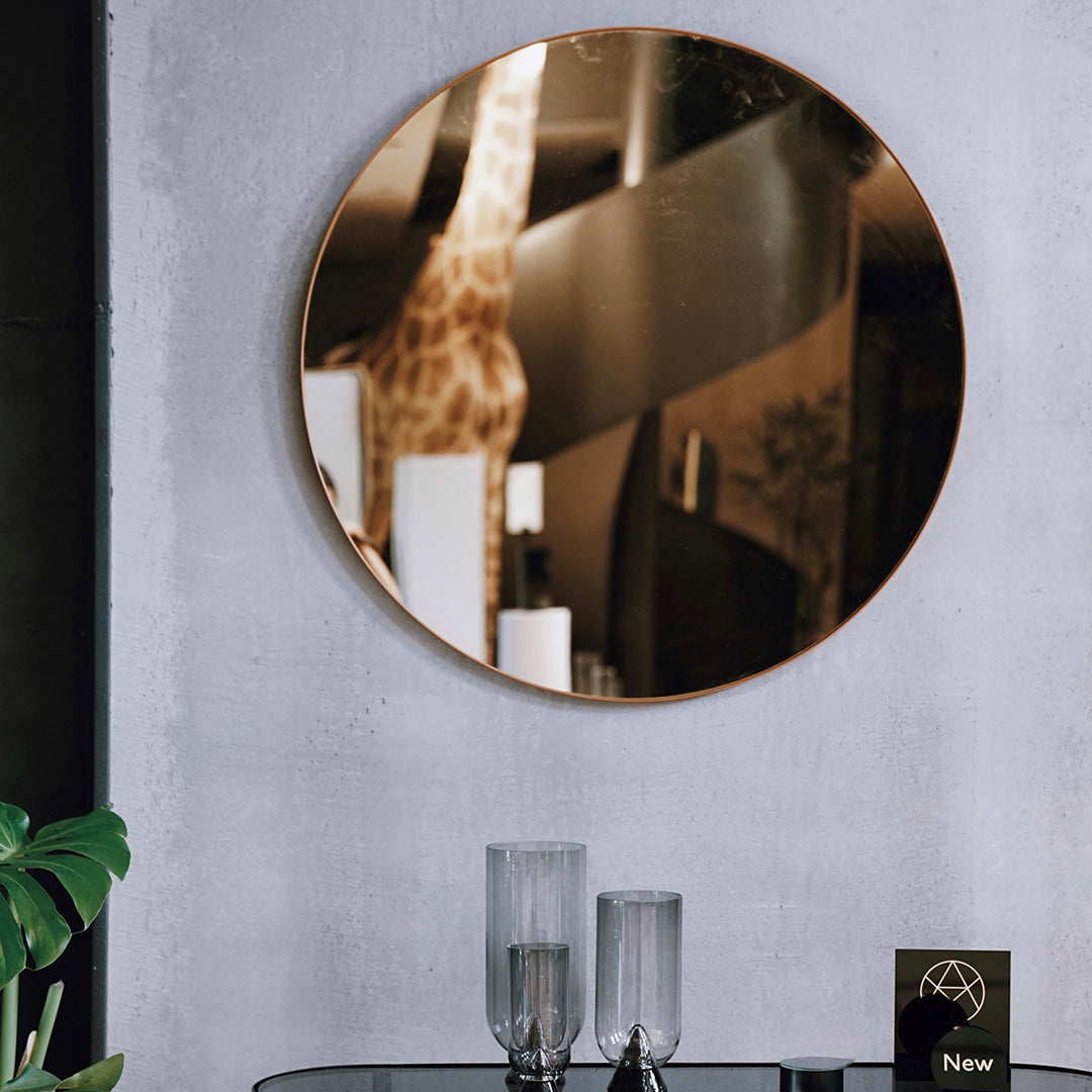 Circular mirror with a sleek copper frame reflecting a modern, minimalist decor featuring warm tones and smooth surfaces.