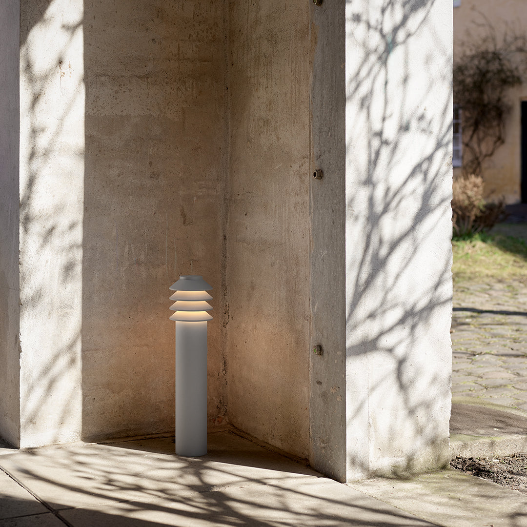 Modern outdoor lamp with a sleek gray base and layered white shade, casting soft light and shadows on stone surfaces.