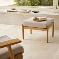 Light gray upholstered ottoman with a natural wood frame, set against a serene, neutral-toned environment.