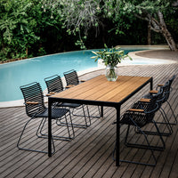 Stylish wooden outdoor table with black and striped chairs, surrounded by lush greenery and a blue pool.