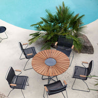 Black and white striped chairs surround a circular wooden table, set against a turquoise pool and lush greenery.