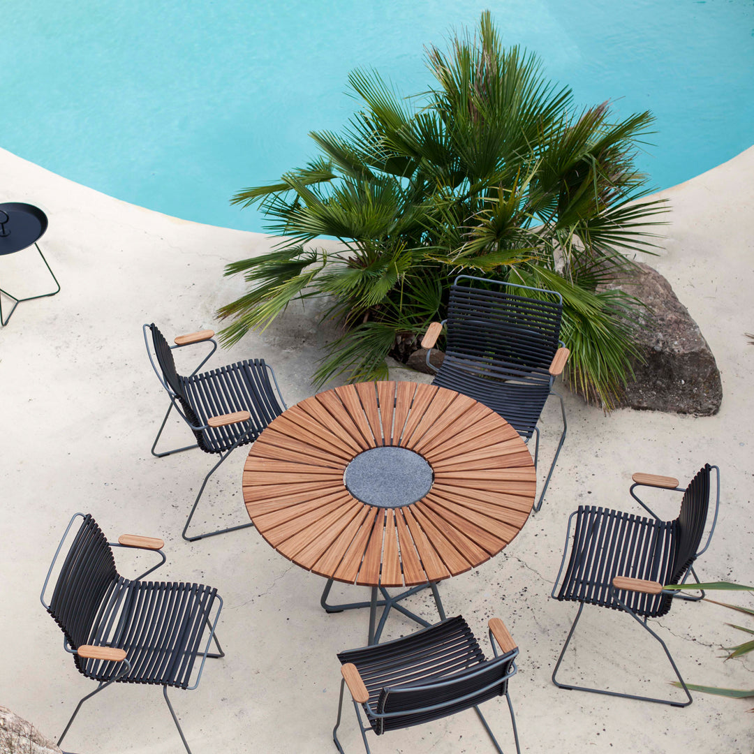 Black and white striped chairs surround a circular wooden table, set against a turquoise pool and lush greenery.