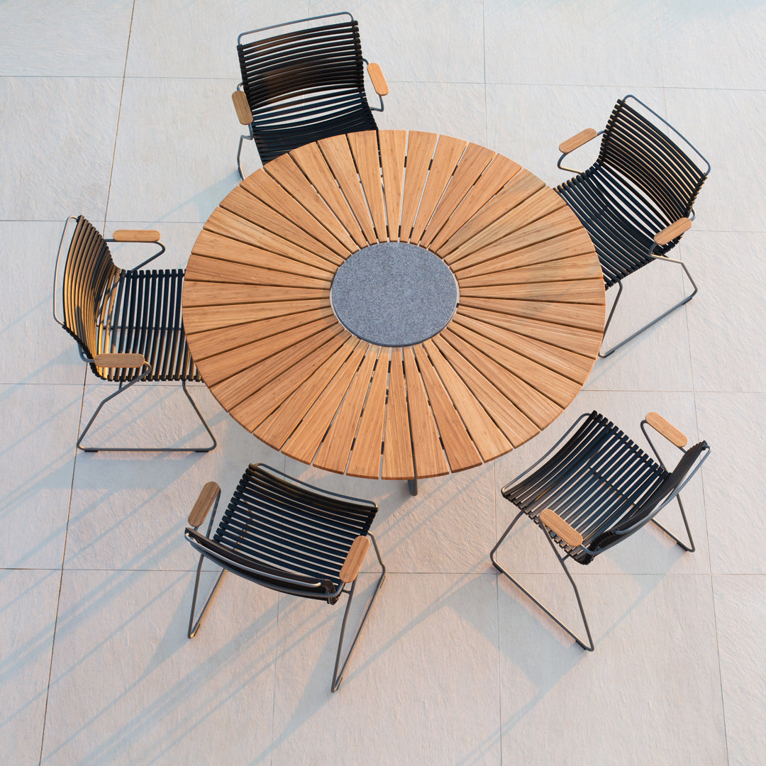 Round wooden dining table with gray center, surrounded by black striped chairs. Elegant outdoor seating design.