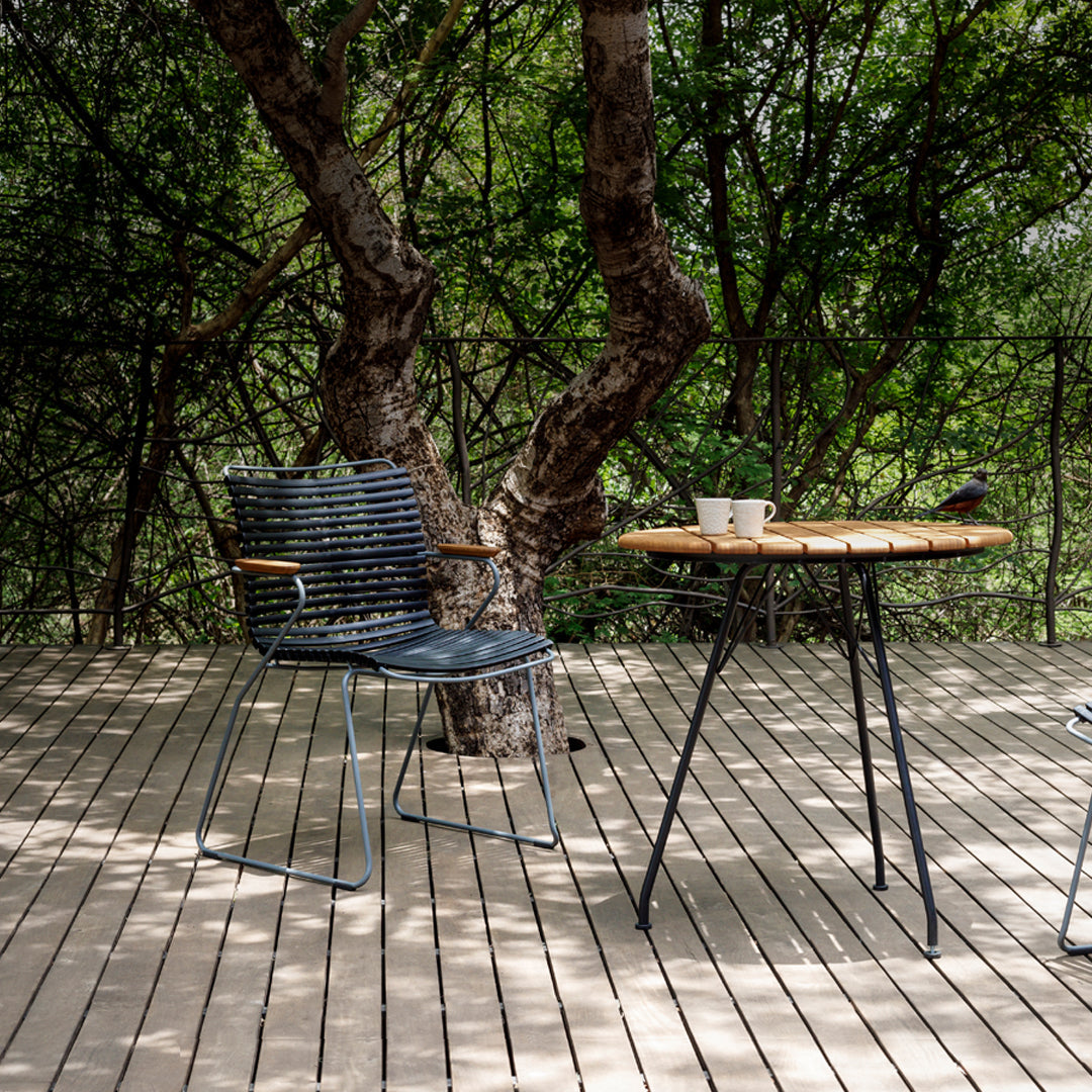"Stylish blue striped chair beside a natural wood table with a dark metal base, set on a wood deck amidst greenery."