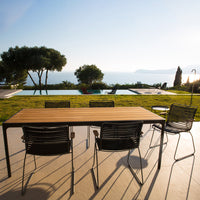 Stylish wooden dining table with black woven chairs, set against a sunny outdoor landscape and blue ocean view.
