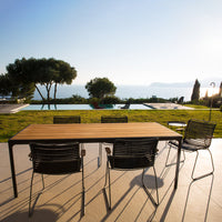 Natural wood table with black metal chairs, set against a serene outdoor landscape with blue skies and lush greenery.