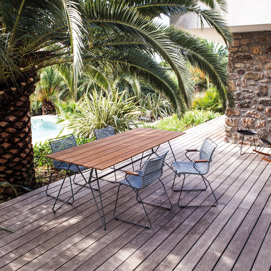 Outdoor dining set with a wooden table and blue metal chairs, surrounded by lush greenery and stone features.
