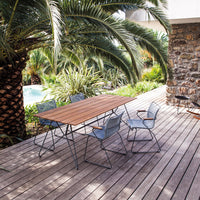 Outdoor dining set with a wooden table and blue metal chairs, surrounded by lush greenery and stone features.