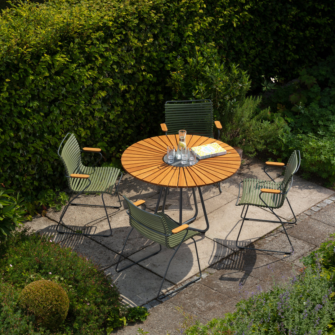 Round wooden patio table with green chairs, set against lush greenery and stone paving. Ideal for outdoor dining.