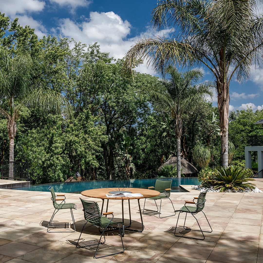 "Round wooden table with green patterned chairs on a stone patio, surrounded by lush greenery and blue skies."