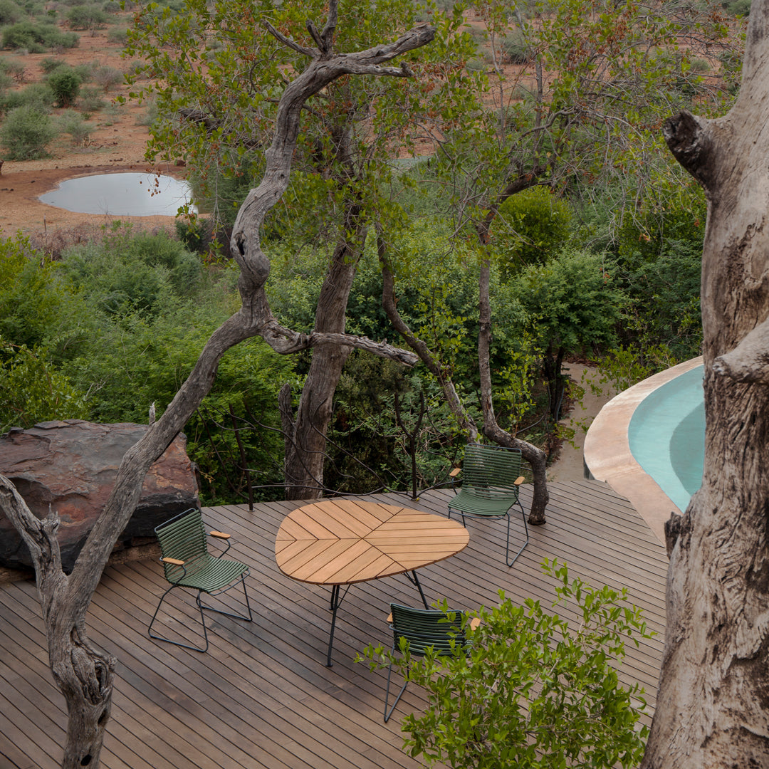 Natural wood table with a circular design, surrounded by green chairs against a backdrop of lush foliage and earthy tones.