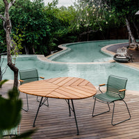 "Round wooden table with green striped chairs beside a curved pool, surrounded by lush greenery."