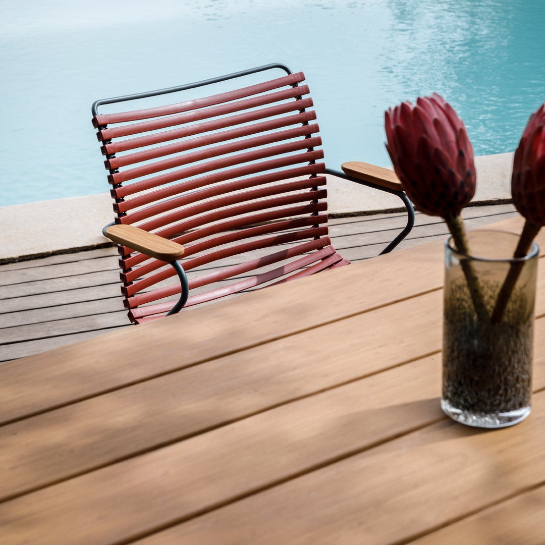 Red striped chair, wooden table, and glass vase with pink flowers against a serene blue pool backdrop.
