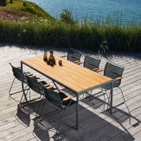 Wooden outdoor dining table with sleek metal frame, surrounded by textured green chairs on a sunny deck.