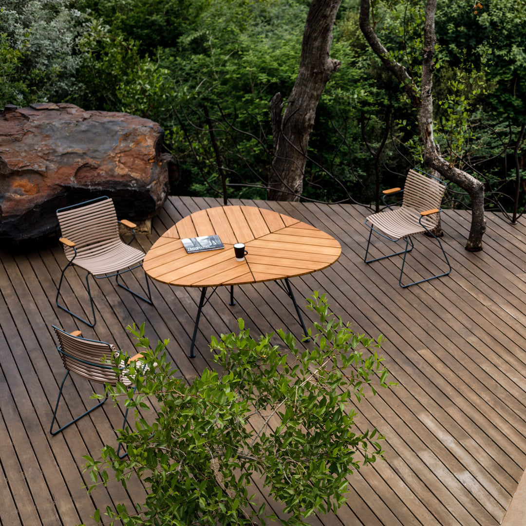 Natural wood tabletop, beige chairs with black legs, surrounded by lush greenery and wooden decking.