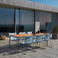 Light blue woven chairs surround a sleek wooden dining table on a natural wood deck, under clear skies.