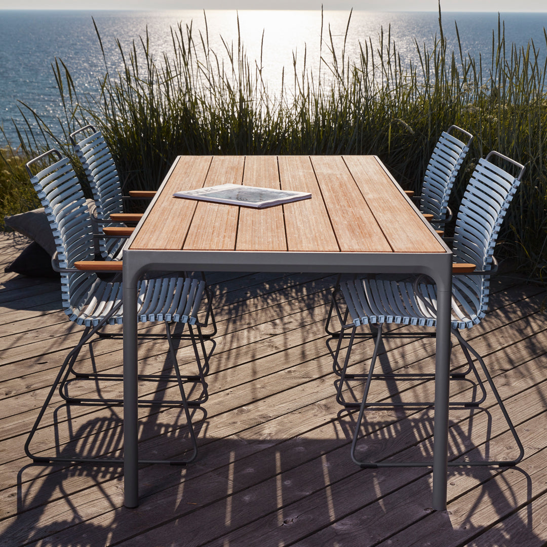 "Modern outdoor dining table with natural wood finish and six blue-striped chairs against a coastal backdrop."