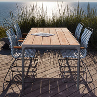 "Modern outdoor dining table with natural wood finish and six blue-striped chairs against a coastal backdrop."