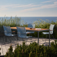 "Outdoor dining set with sleek blue and silver striped chairs and a natural wood tabletop, set against a serene coastal backdrop."