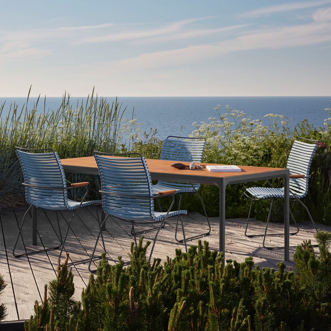 "Outdoor dining set with sleek blue and silver striped chairs and a natural wood tabletop, set against a serene coastal backdrop."