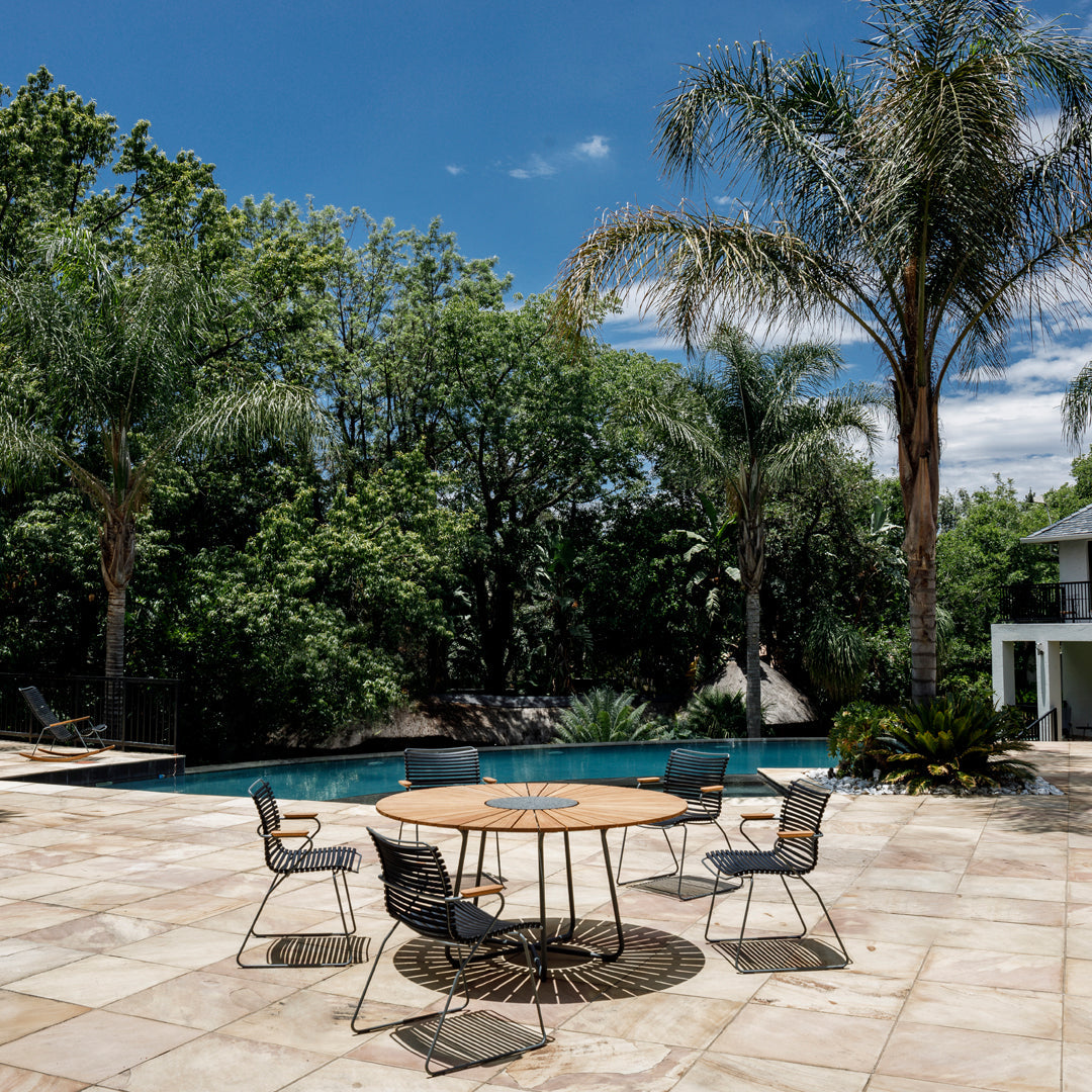 "Round wooden table surrounded by black chairs on a stone patio with green palms and blue sky."
