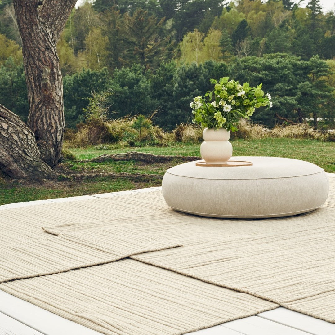 Beige round pouf on a textured neutral rug, surrounded by greenery and a simple vase of flowers.