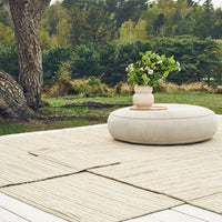 Beige round pouf on a textured neutral rug, surrounded by greenery and a simple vase of flowers.