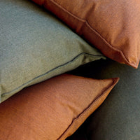 Stack of textured throw pillows in olive green and burnt orange, featuring subtle patterns and contrasting edges.