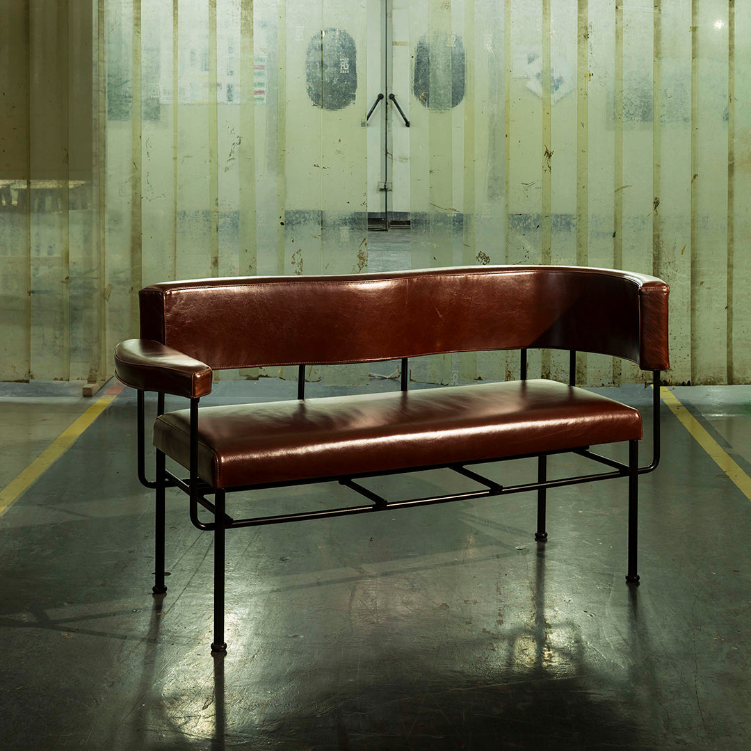 Brown leather sofa with a sleek black metal frame and minimalist design, set against a textured industrial background.