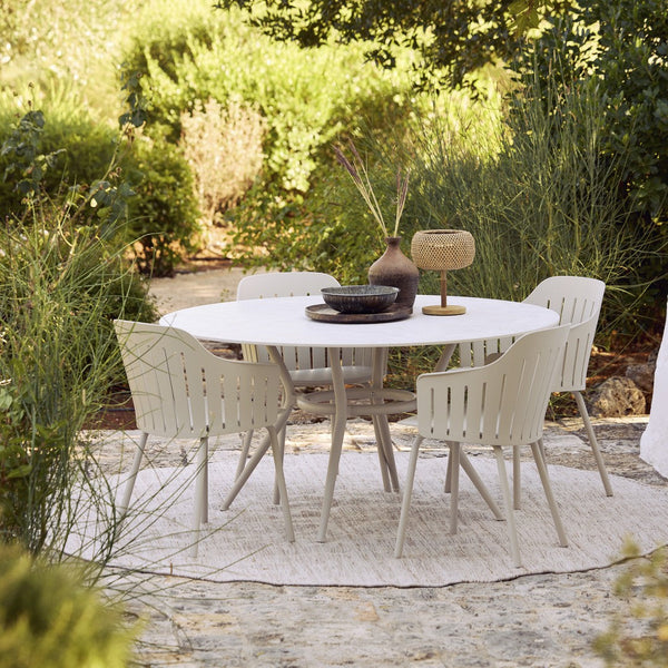 Round white table with beige chairs, set on a textured neutral rug in a lush green garden setting.