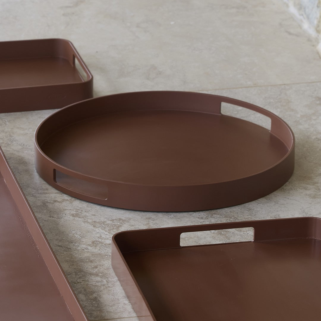 Round brown tray with smooth surface and cut-out handles, paired with rectangular brown trays on a light marble background.