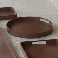 Round brown tray with smooth surface and cut-out handles, paired with rectangular brown trays on a light marble background.