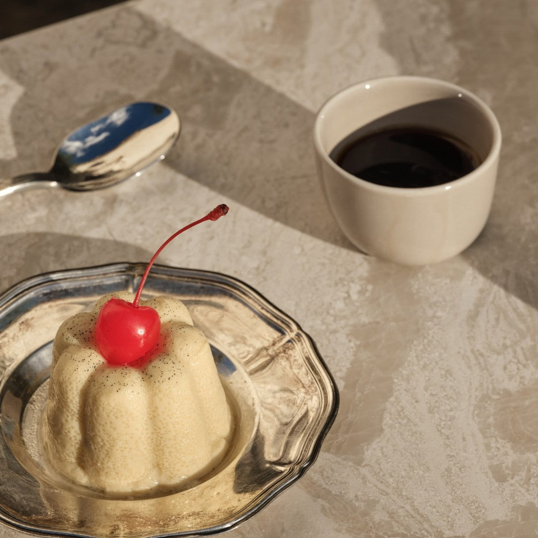 Yellow dessert with a red cherry on top, served on a silver plate, alongside a white cup of dark liquid.
