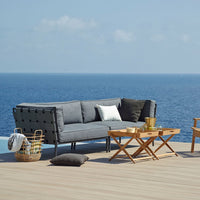 Stylish gray outdoor sofa with black cushions, paired with wooden tables and neutral accessories against a blue ocean backdrop.