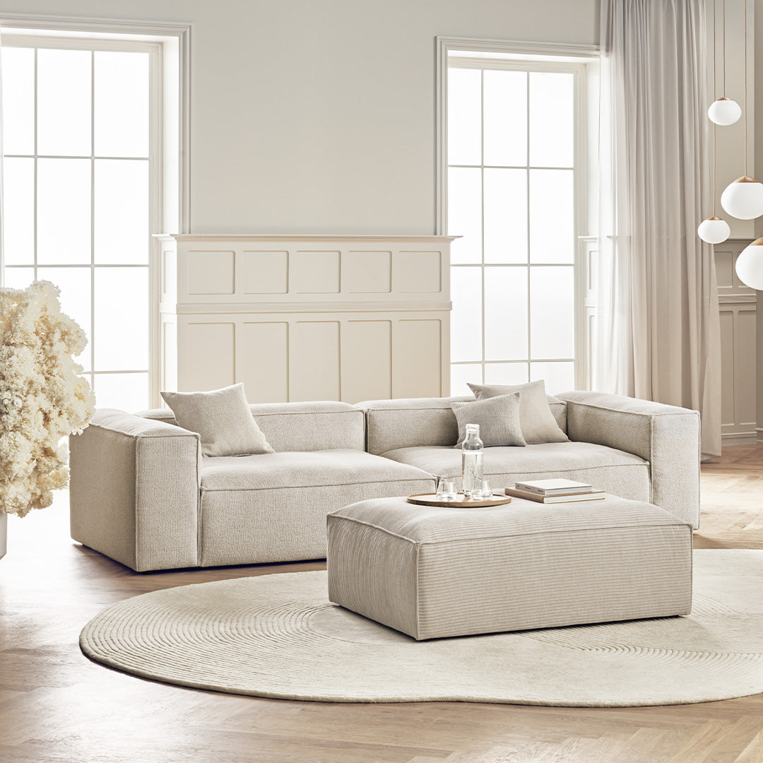 Light beige modern sectional sofa with coordinated ottoman on a soft cream circular rug in a bright, airy room.