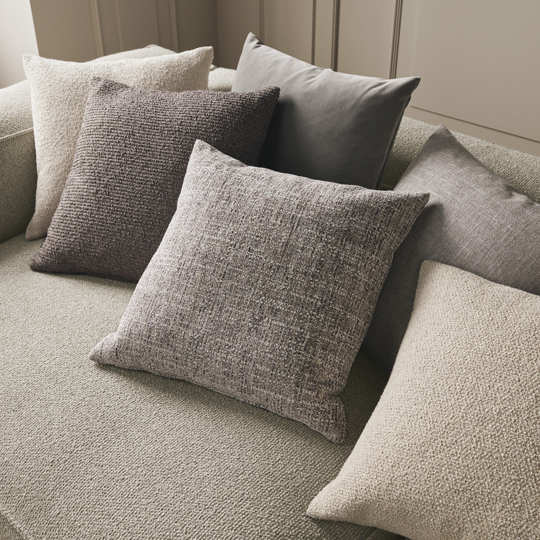 Assorted decorative pillows in soft greys, taupes, and textured patterns, arranged on a plush green sofa.