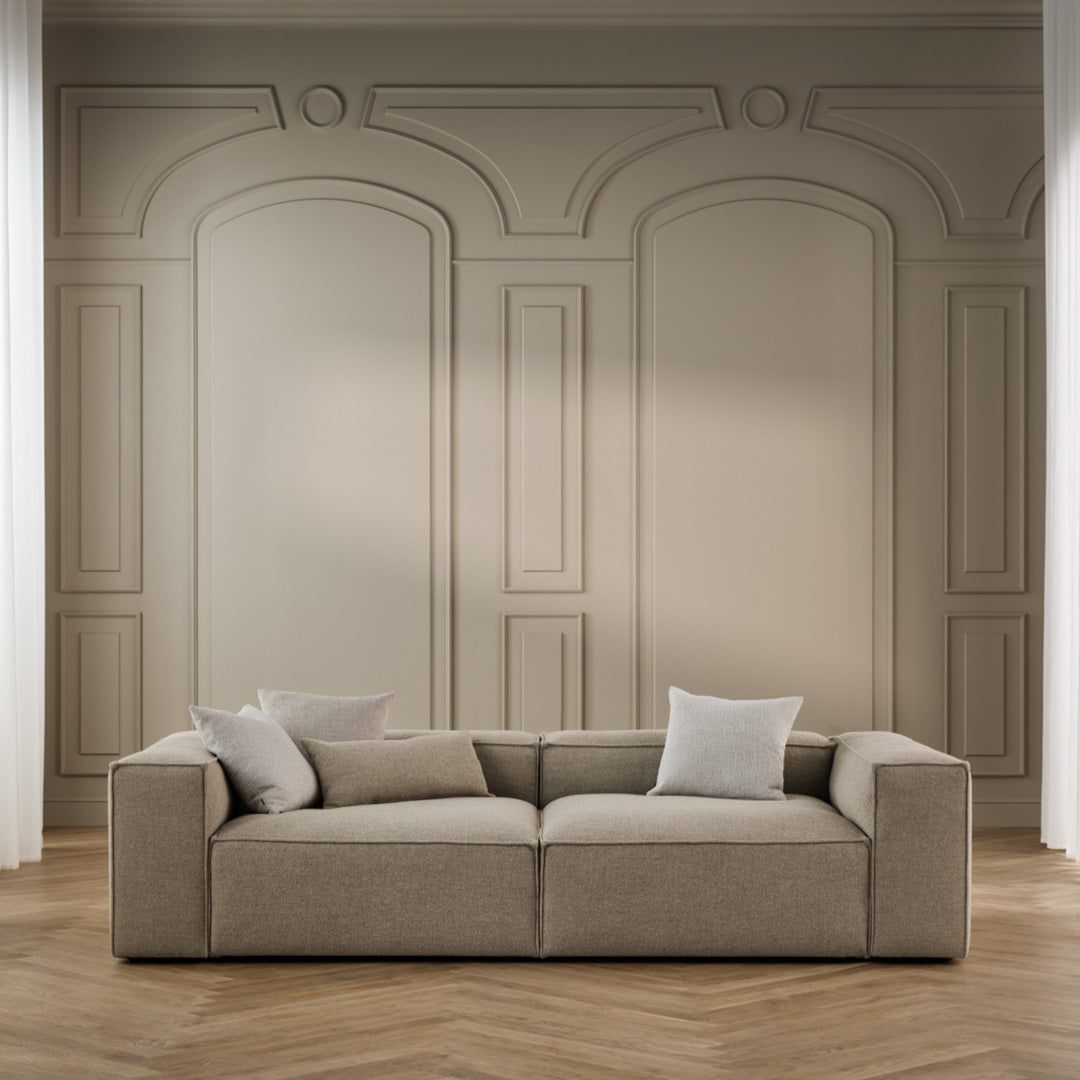 Beige sectional sofa with light gray cushions against a neutral patterned wall and wooden herringbone floor.
