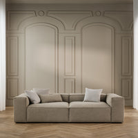 Beige sectional sofa with light gray cushions against a neutral patterned wall and wooden herringbone floor.