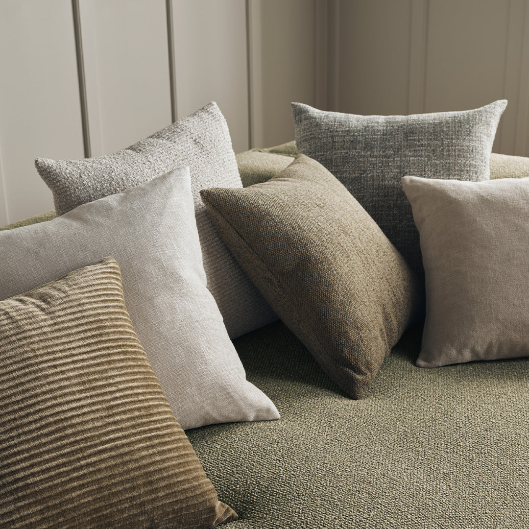 Assorted decorative pillows in muted tones of beige, gray, and earthy patterns on a soft green background.