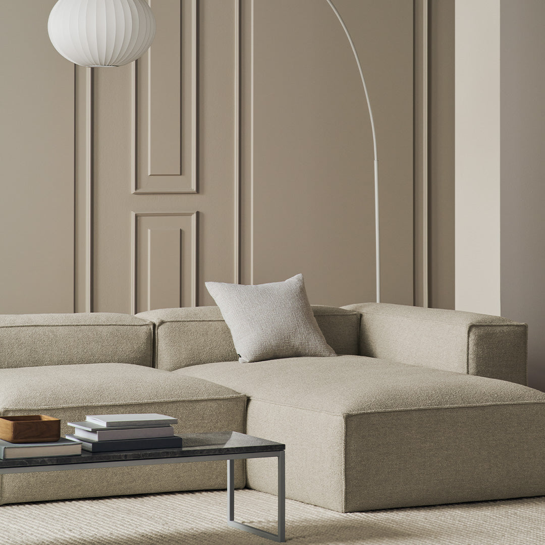 Beige sectional sofa with subtle texture, paired with a minimalist white pillow and sleek gray coffee table in a neutral setting.