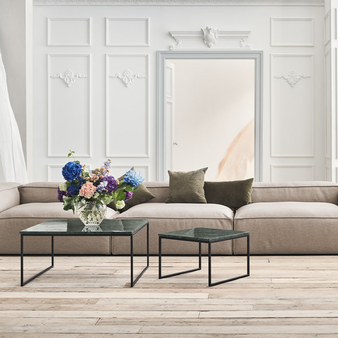 Beige sofa with dark green pillows, clear glass tables with black metal legs, and vibrant floral arrangement.