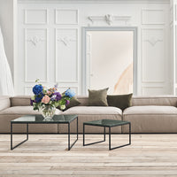 Beige sofa with dark green pillows, clear glass tables with black metal legs, and vibrant floral arrangement.