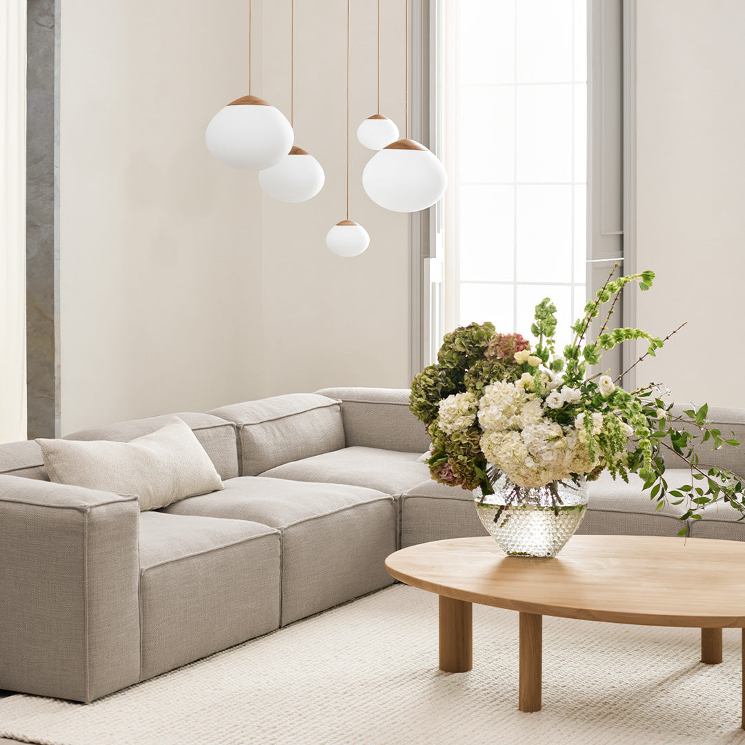 Beige sectional sofa with neutral tones, white pendant lights, and a vibrant floral arrangement on a light wood coffee table.