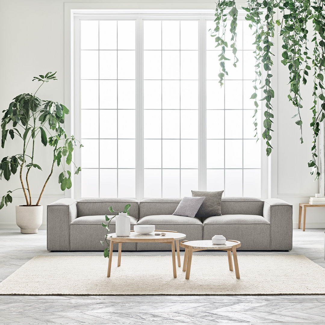 Modern grey sectional sofa in a bright, airy room with natural light and greenery, accented by neutral-toned rugs and decor.