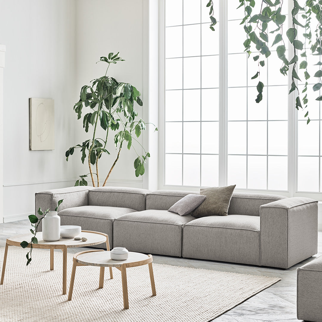Modern light gray sectional sofa with subtle textured pattern, set in a bright, airy room with greenery.