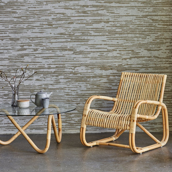Rattan chair with a natural finish, paired with a glass top table, against a textured beige wall.