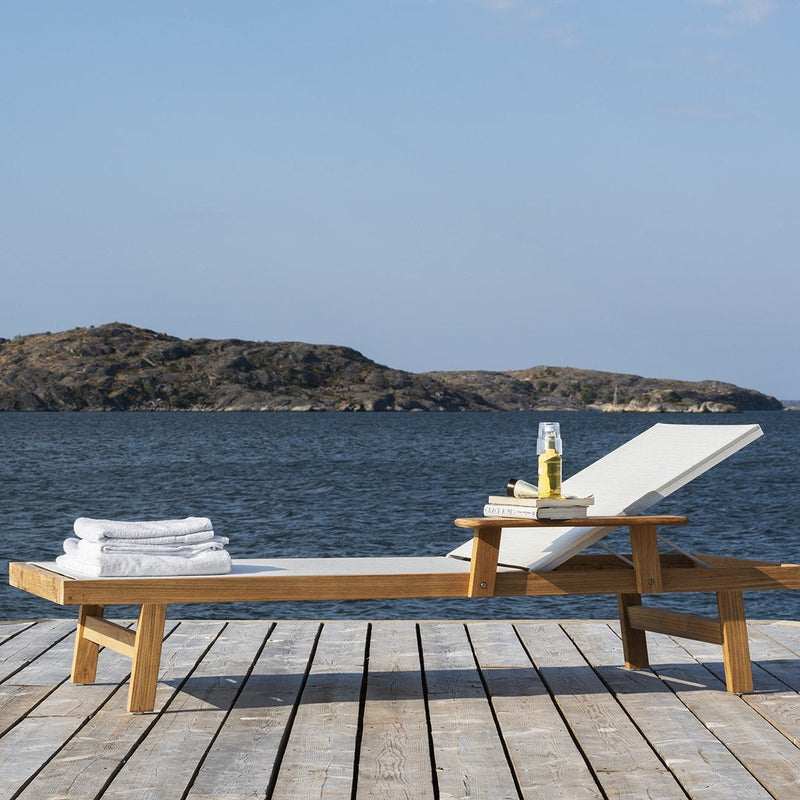 "White chaise lounge with natural wood frame, set against a blue water backdrop under a clear sky."