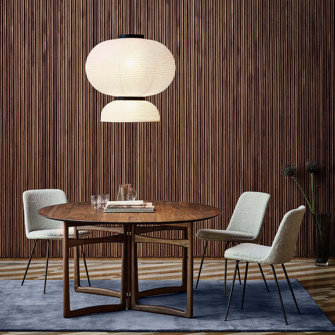 Round wooden table with beige chairs on a blue rug, complemented by a soft white pendant light and wooden backdrop.